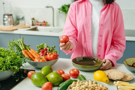 Woman in pink overshirt holds tomato in kitchen with fresh fruits and veggies.